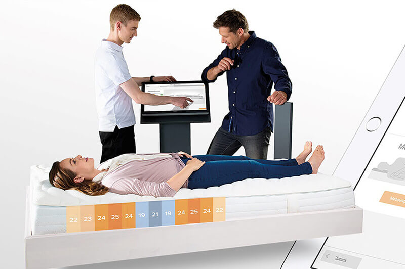 Frau liegt auf Matratze, auf der Festigkeitszonen markiert sind, Zwei Schlafexperten besprechen sich an Monitor